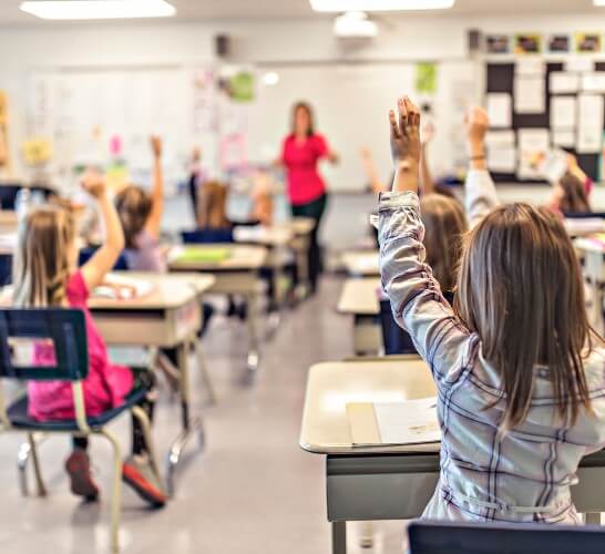 A girl facing the front of a classroom with her hand up